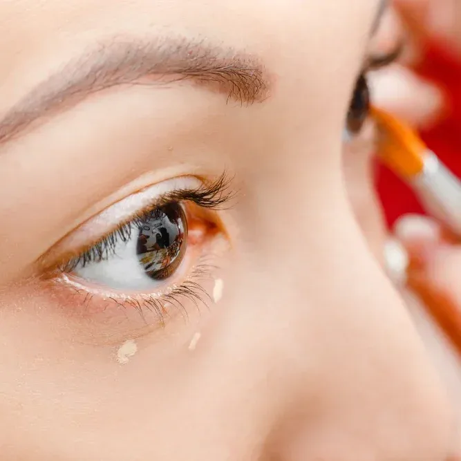 Woman applying oil-free foundation around eye area with makeup brush, preparing smooth base for special event makeup look