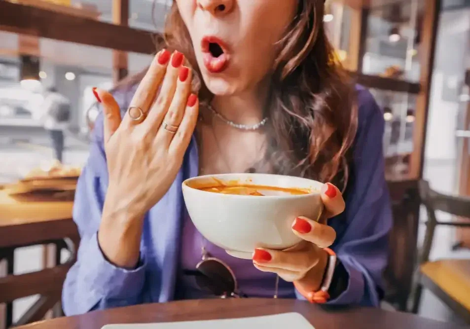 A woman reacts with surprise while eating spicy soup, illustrating the types of irritating foods to avoid on the first day of lip tattoo healing.