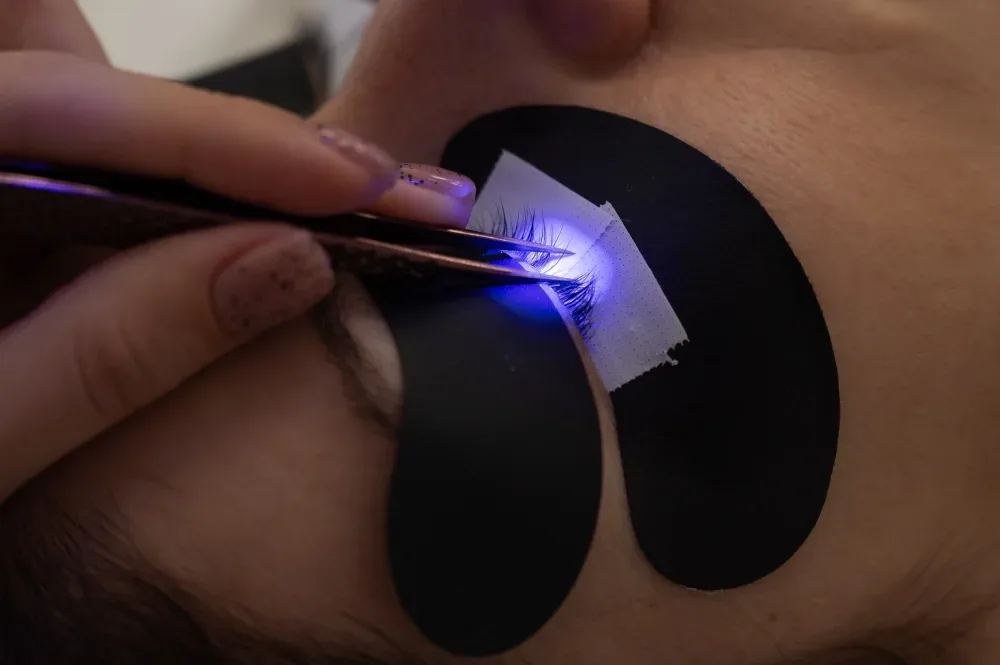 Beautician uses UV/LED curing tool with protective patches on client's closed eyes during safe extension bonding