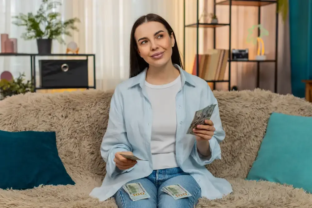 A woman holding cash sits on a couch, planning her budget for eyelash extension maintenance and fills.