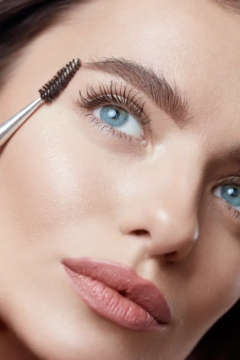 A woman's eyebrow is groomed with a spoolie, representing the various aftercare products needed for lash maintenance.