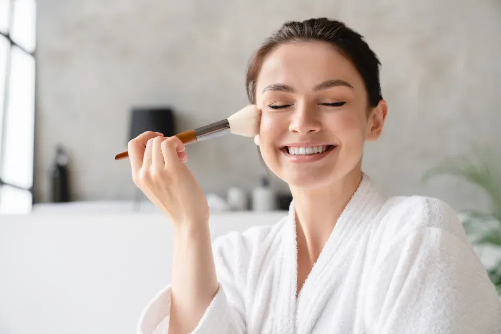 A smiling woman in a robe happily applies makeup, symbolizing the confidence that comes from trusting lash facts.