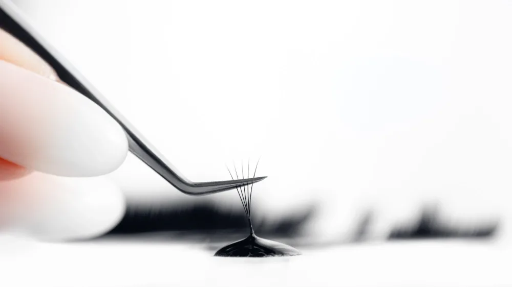 A lash fan is dipped into adhesive with tweezers, preparing for a patch test to minimize allergic reaction risks.