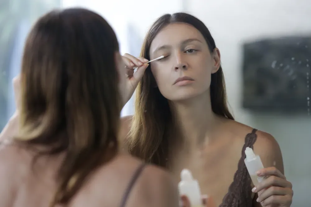 A woman carefully applies eyelash serum while looking in a mirror, representing making a conscious choice for lash health and beauty.