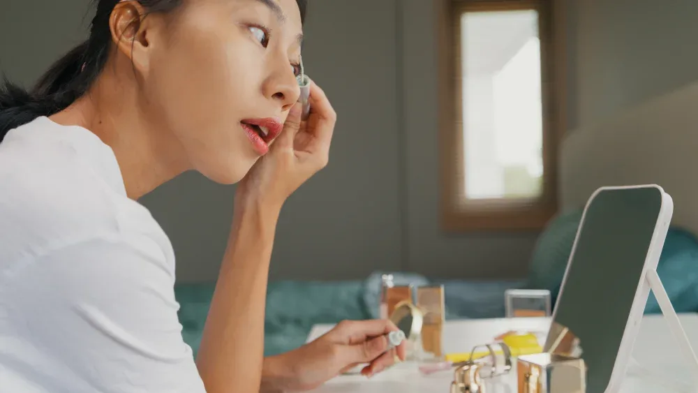 A woman looks in a mirror while correctly applying eyelash serum along her lash line, demonstrating the proper application technique.