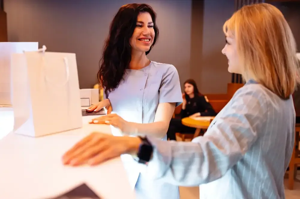 A client at a salon reception desk discusses changing her lash style for more volume with her technician.
