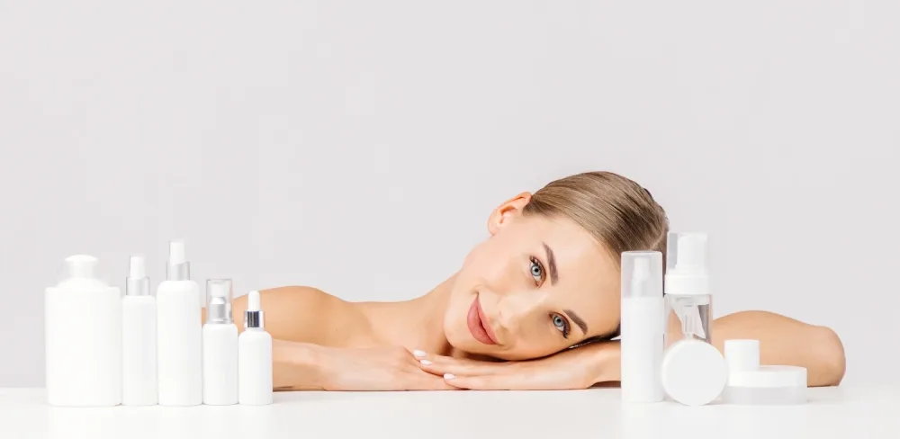 A smiling woman rests among a collection of cosmetic bottles, representing the need to budget for aftercare products after a lip tattoo.