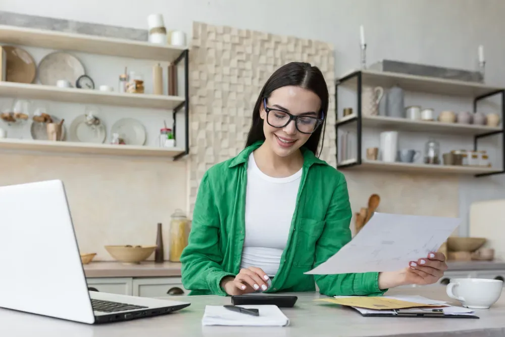 A woman in a green shirt at her desk with a laptop and papers, researching and calculating the average cost of lip tattooing.