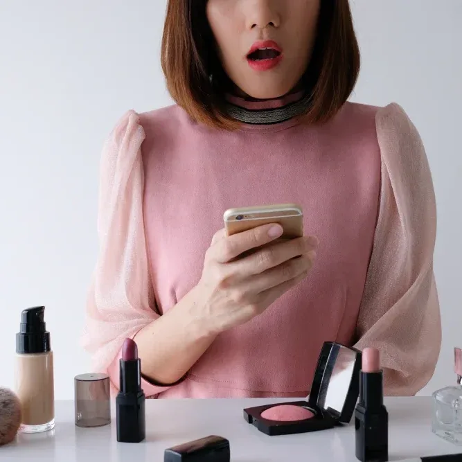A woman looks at her phone with surprise, surrounded by cosmetics, representing the high initial cost of a tattoo versus filler maintenance.